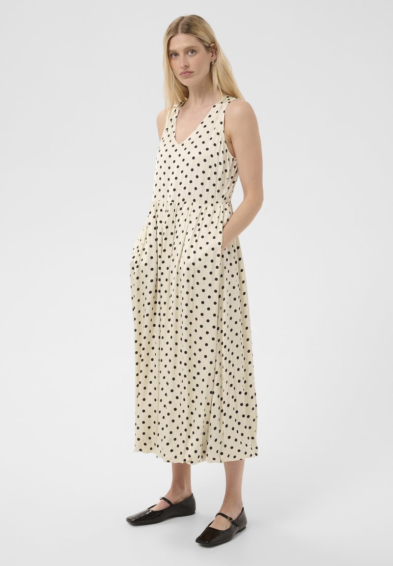 Femme aux cheveux blonds portant une robe crème sans manches à pois noirs et des chaussures plates noires sur un fond gris uni.