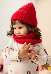 Cappello di lana a costine rosso e sciarpa abbinata su un bambino. Maglietta a maniche lunghe color crema con motivi festivi. Texture morbida, colore vivace, aspetto accogliente.