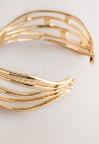 Gold cuff bracelet featuring wavy, elongated shapes with a polished finish. The design includes open spaces between the curved sections.