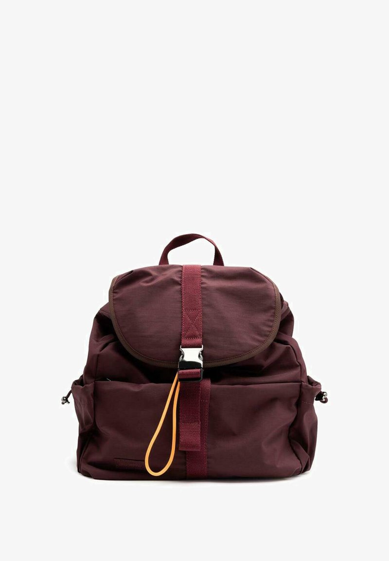 Mochila de tela color marrón con una solapa redondeada, correas de hombro ajustables, una hebilla plateada y una pestaña de tirón naranja en contraste. Cuenta con bolsillos en la parte frontal.