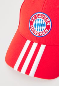 Red baseball cap with embroidered FC Bayern München logo. Features three white stripes on the visor. Made of textured fabric.