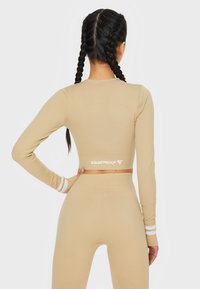 Beige Langarm-Crop-Top mit weißen Streifenakzenten und "SQUATPROOF"-Logo auf dem Rücken; figurschmeichelnder Stoff, glatte Textur.