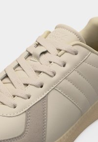 Κοντινή λήψη σε μπεζ αθλητικό παπούτσι με υφασμένες κορδόνια, ραμμένα πάνελ και καουτσούκ σόλα, που αναδεικνύει τη γλώσσα και τις λεπτομέρειες του άνω μέρους.