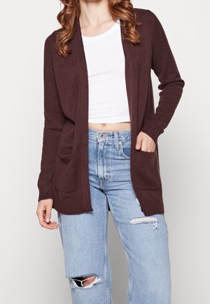 Femme aux cheveux ondulés porte un cardigan marron ouvert sur un top court blanc et un jean bleu clair déchiré, une main dans la poche du cardigan.
