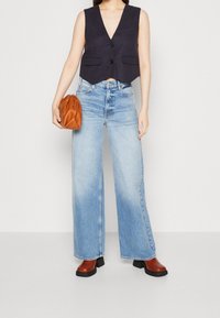 Marineblaues Leinen-Ärmelwesten mit zwei vorderen Taschen, hochgeschnittene hellblaue weit geschnittene Jeans, braune Lederhandtasche und braune Stiefel mit Absatz.