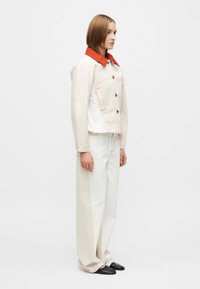 Μπεζ βαμβακερό μπουφάν με κόκκινο γιακά, πέντε κουμπιά και τσέπη στο στήθος. Συνδυασμένο με φαρδιές λευκές παντελόνες με αντίθετες πλαϊνές λωρίδες.