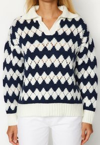 Donna che indossa un maglione a maglia con motivo a diamante blu navy e bianco, con colletto bianco e pantaloni bianchi, mostrata dalla collo ai fianchi.