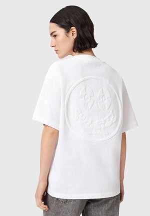 Kvinde med kort mørkt hår iført en hvid oversized T-shirt med et præget smiley-ansigtsdesign på ryggen og grå jeans.