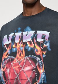 Μαύρο βαμβακερό μπλουζάκι με μεγάλο, πολύχρωμο λογότυπο "NIKE" με γραφικά φλόγας και στυλι ζαρισμένο μπάσκετ που είναι διακοσμημένο με αλυσίδες.