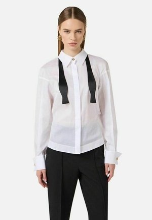 Donna che indossa una camicia bianca a maniche lunghe con colletto a fiocco nero sbottonato e pantaloni neri su misura, in piedi davanti a uno sfondo semplice.