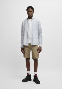 Camicia a maniche lunghe a righe azzurre e bianche, indossata sopra una maglietta bianca, abbinata a pantaloni cargo color sabbia e sneaker alte nere.