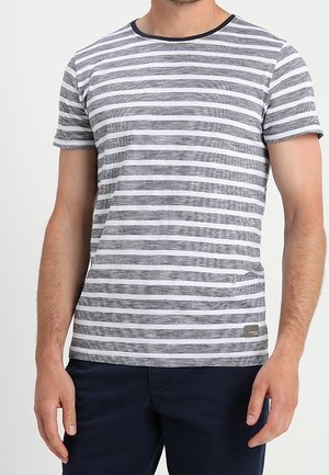 Mand iført en grå og hvid t-shirt med horisontale striber og mørkeblå bukser, stående foran en ensfarvet baggrund.
