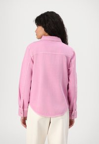 Pink denim button-up shirt with a curved hem, long sleeves, and visible stitching details. The fabric appears textured and lightweight.