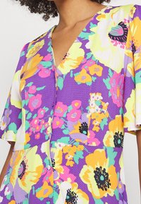 Abito floreale colorato con base viola e accenti gialli, rosa e verdi. Design a V, maniche corte, tessuto strutturato.