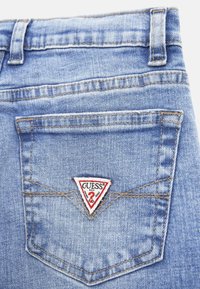 Vaqueros de mezclilla azul claro con un bolsillo trasero que presenta un parche de logo en forma de triángulo en rojo y blanco de "Guess" y detalles de costura en contraste.