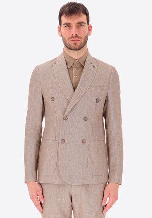 Uomo che indossa una giacca doppiopetto beige sopra una camicia marrone con bottoni, in piedi davanti a uno sfondo chiaro e uniforme.