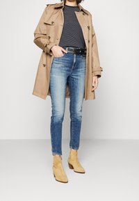 Béžový trenčkot přes námořnicky modrý a bílý pruhovaný top, modré slim-fit džíny a hnědé semišové kotníkové boty na dřevěném podpatku. Jednoduchý pásek s přezkou.