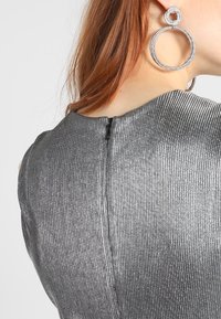 Top argent à côtes avec un col montant et un détail à fermeture éclair au dos. De grandes boucles d'oreilles circulaires complètent le look. Texture lisse.