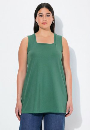 Joven mujer con cabello largo y oscuro que lleva una camiseta sin mangas verde con escote cuadrado y vaqueros azules, de pie frente a un fondo blanco.