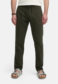Olivenfarvede teksturerede bukser med elastisk talje og snøring, der har sidelommer og er parret med grå sandaler. Stofvet har et subtilt mønster.