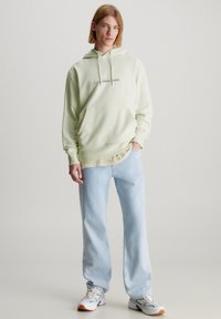 Felpa verde chiaro con tasca frontale e logo, indossata sopra una maglietta crema. Abbinata a jeans azzurri chiari e sneakers bianche con accenti colorati.