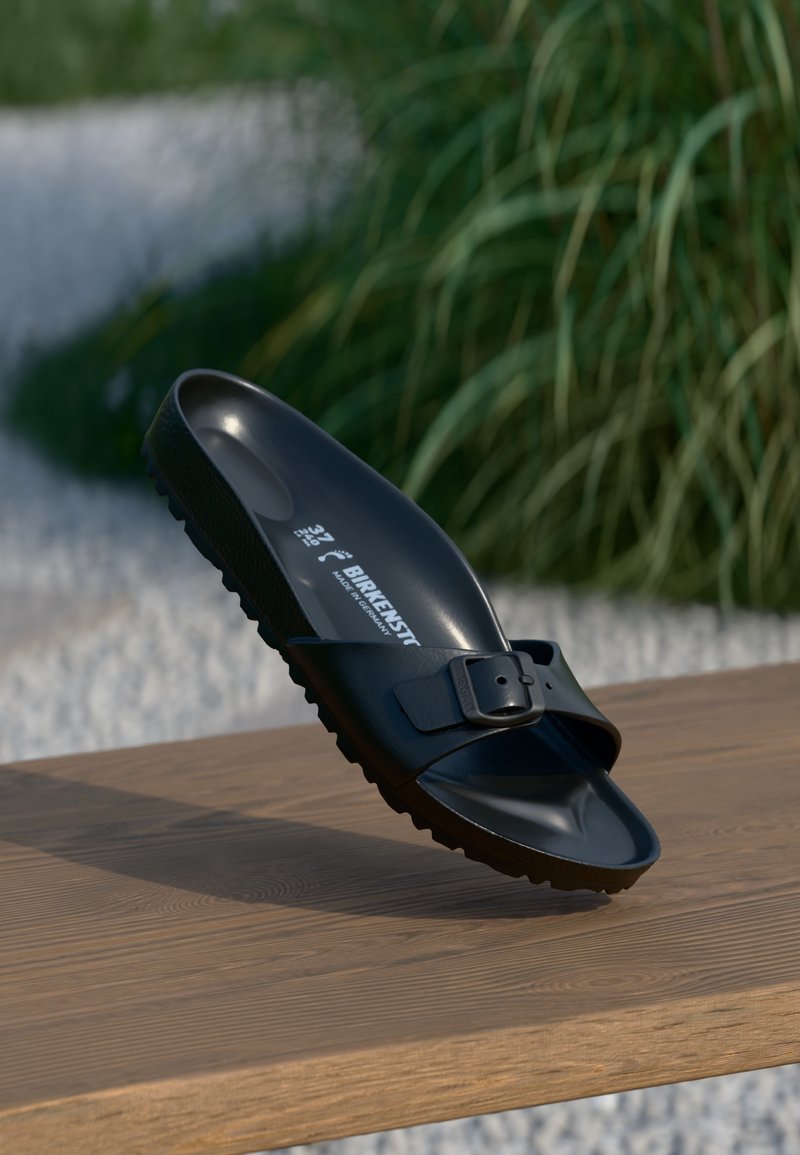 Sandale Birkenstock noire à enfiler avec boucle réglable reposant sur une surface en bois, avec un arrière-plan flou de verdure et de gravier blanc.