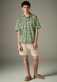 Chemise à manches courtes verte à motifs géométriques, associée à un short beige clair texturé et des chaussures tressées marron clair. Fond neutre.