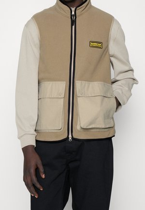 Man draagt een beige en lichtbruine vest met ritssluiting, met twee voorzakken met klep en een gele "Barbour International" logo patch op de borst over een licht langärmelig shirt.