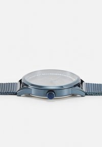 Une montre métallique bleue avec un boîtier rond, un verre en face et un bracelet en maille. Elle présente une couronne bleue, un design épuré et un cadran minimaliste.