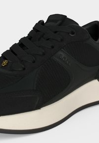 Baskets noires à lacets avec panneaux latéraux texturés et semelle blanche, ornées d’un petit emblème doré sur les lacets et d’un marquage en relief "BOSS".