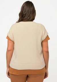Beige pletený svetr s krátkými rukávy, s ribbovanou texturou a volným střihem. Na sobě má hnědé tričko s vyzývavým výstřihem, s viditelnými bočními kapsami.