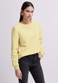 Giovane donna che indossa un maglione giallo pallido a trama e jeans neri, in piedi con una mano in tasca contro uno sfondo neutro.