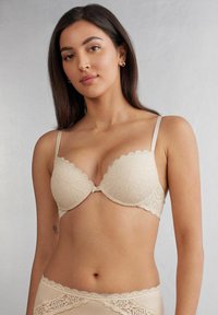 Beige spets-bh med skulpterad kant, släta kupor och tunna justerbara band. Dekorativt hårdvara i mitten fram.