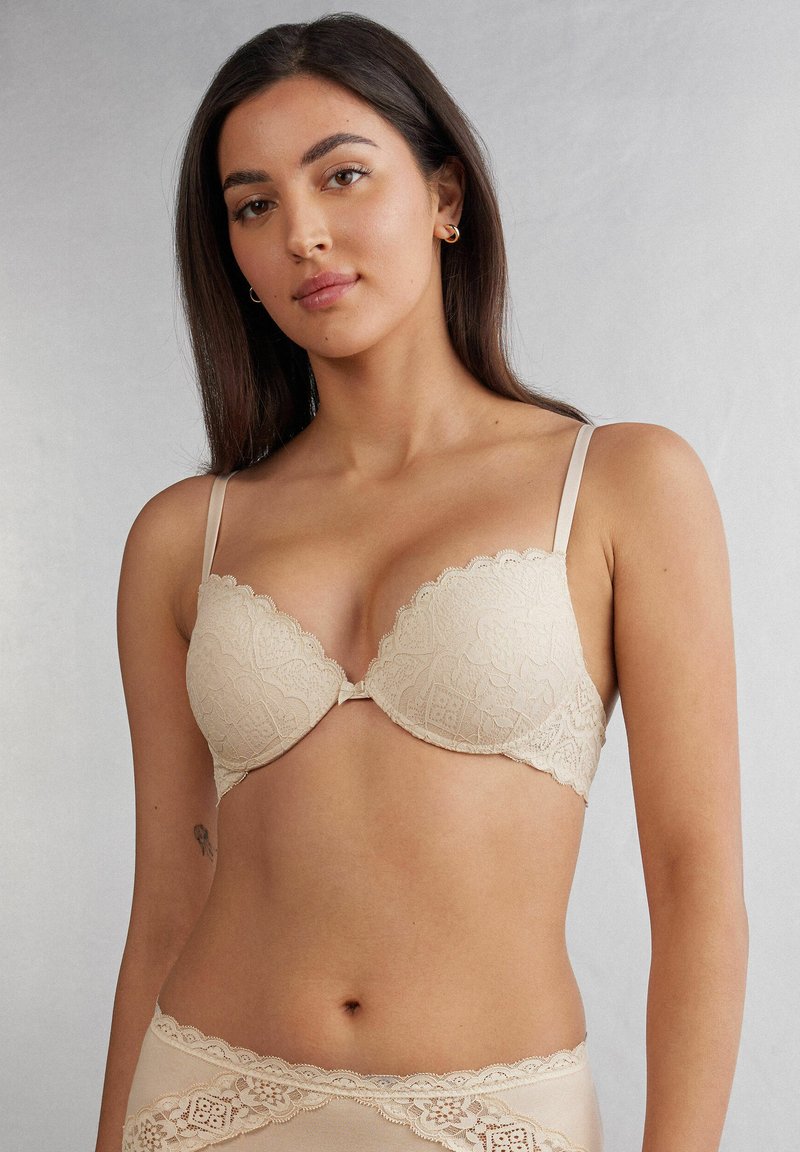 Beige spets-bh med skulpterad kant, släta kupor och tunna justerbara band. Dekorativt hårdvara i mitten fram.