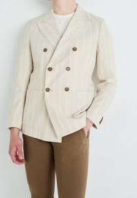 Béžový lněný dvouřadý blazer s bílými pruhy, s hnědými knoflíky a dvěma předními kapsami. V páru s hnědými kalhotami.
