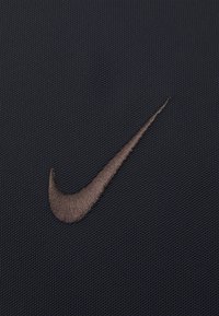 Logo Nike realizat din fir de bronz, brodat pe un material texturat de culoare neagră, având un design neted și curbat pe fundal.