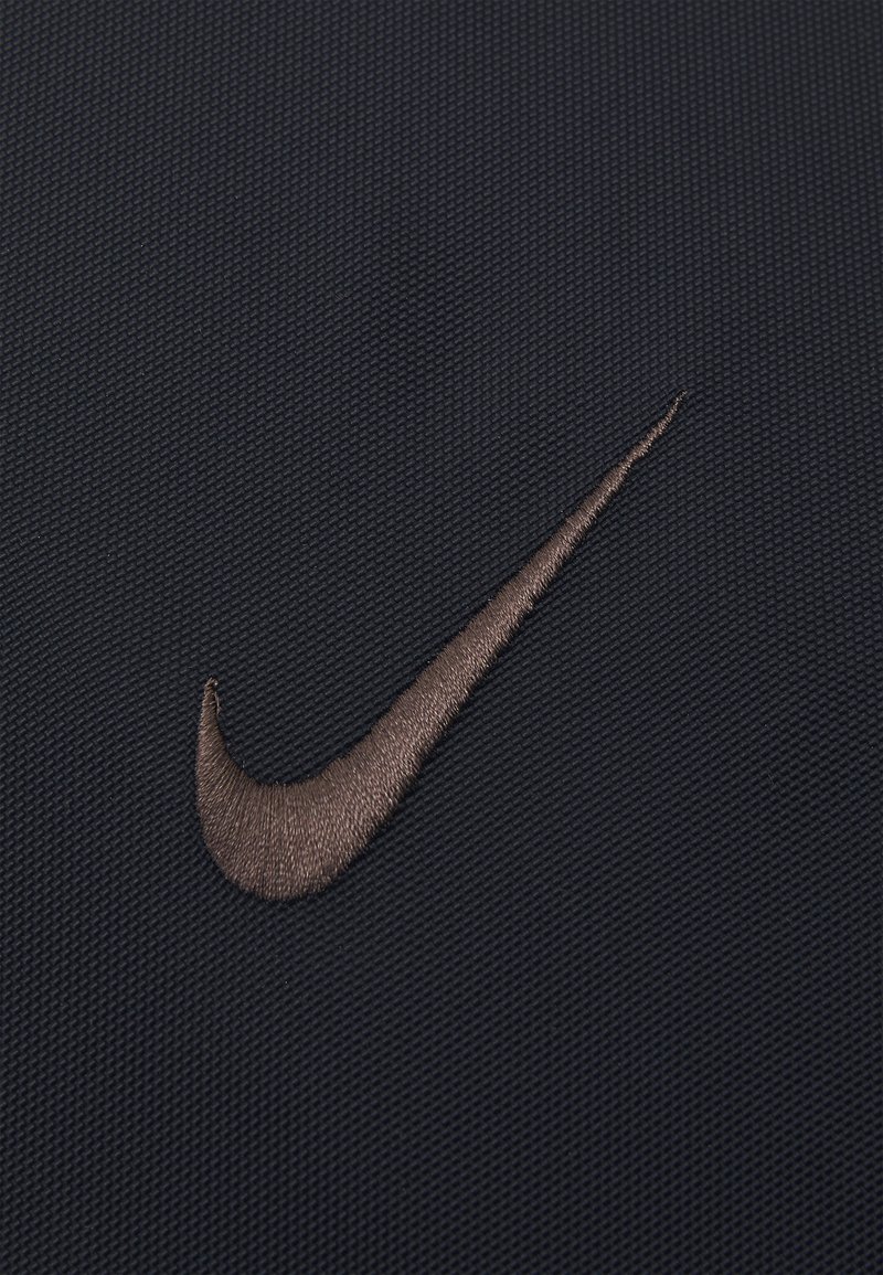 Logo Nike en fil de bronze, brodé sur un tissu noir texturé, présentant un design lisse et courbé sur fond.