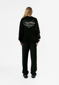 Sudadera negra de gran tamaño con texto blanco en la parte de atrás, combinada con pantalones negros holgados y calzado negro grueso.