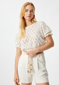Top blanco de punto con patrón ondulado, mangas cortas abullonadas, combinado con pantalones cortos beige que cuentan con un cinturón trenzado y cuentas decorativas.