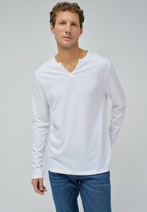 Camisa blanca de manga larga con escote en V y tapeta de tres botones. Confeccionada en una tela suave y ligera, tiene un corte ajustado. Combinada con jeans azul.