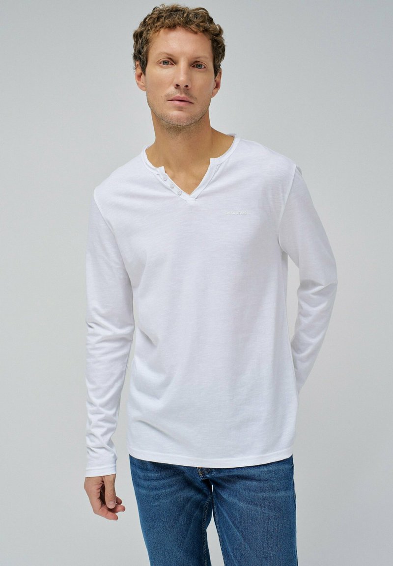 Weißes Langarmhemd mit V-Ausschnitt und drei Knöpfen. Aus weichem, leichtem Stoff und in schlanker Passform. Kombiniert mit blauen Jeans.