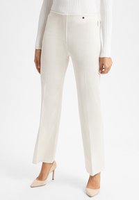 Vrouw in off-white rechte broek en bijpassende beige hoge hakken, met een geribbelde witte lange mouwtop.