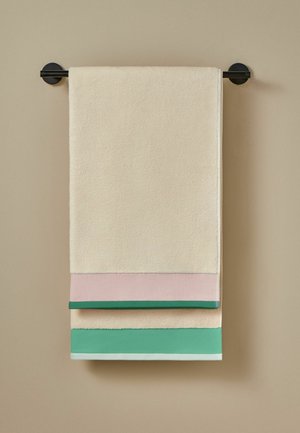 Toalla beige con bordes en bloques de color rosa y verde colgada en una barra para toallas negra montada en la pared frente a una pared marrón claro.