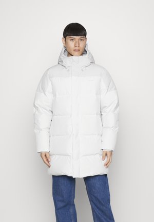 Fehér pufferdzseki kapucnival, steppelt mintával, elöl cipzárral és rugalmas mandzsettákkal. Kék farmernadrággal párosítva.