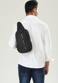 Borsa a tracolla nera realizzata in materiale leggero, con una forma curva, zip frontale e tracolla regolabile, indossata sulla spalla.