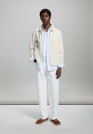 Jeune homme portant un pantalon blanc, une chemise bleu clair, une veste crème avec des lunettes de soleil suspendues, et des sandales marron, debout devant un fond gris uni.