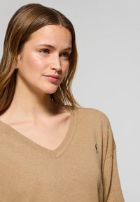 Beige v-hals sweater i blødt strikket materiale, med løs pasform og et lille sort logo på venstre side af brystet.