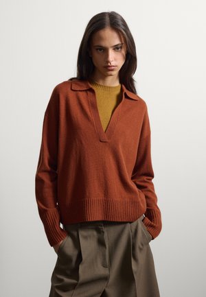 Donna con lunghi capelli scuri indossa un maglione color ruggine sopra una canotta color senape e pantaloni verde oliva, posando con le mani nelle tasche su uno sfondo semplice.