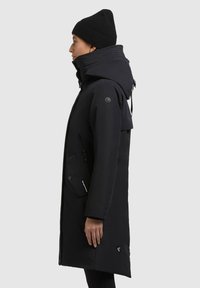 Fekete, hosszú kabát kapucnival, texturált gallérral és cipzáras zsebekkel. Simább anyagból készült, aerodinamikus sziluettel.