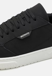 Baskets noires avec une tige lisse, œillets renforcés, semelle en caoutchouc blanc texturé et logo "MORATO" sur le côté.